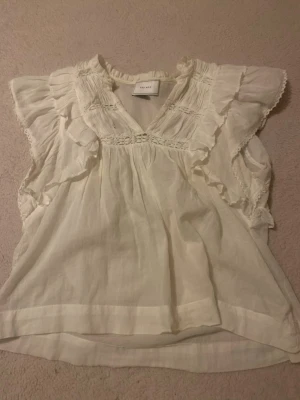 Vit volangblus från neo noir - Söt vit blus från H&M med volanger och spetsdetaljer vid axlar och bröst. Blusen har v-ringning och är tillverkad i ett tunt, luftigt tyg som känns som bomull eller linne. Perfekt för en romantisk och bohemisk stil.