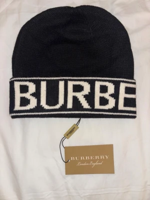Svart Burberry mössa med logga - Snygg svart stickad mössa från Burberry med bred vit kant och stor kontrasterande logotyp runtom. Klassisk beanie-modell som är perfekt för kalla dagar. Mössan är i mjukt material och har en modern, streetinspirerad look.