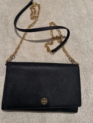 Svart kuvertväska Tory Burch - Stilren svart kuvertväska från Tory Burch i läder med guldfärgad kedjerem och logga framtill. Väskan har flera fack och kortplatser på insidan, perfekt för att hålla ordning på det viktigaste. Klassisk och elegant design som passar till många looks.