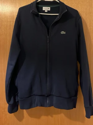 Marinblå zip hoodie från Lacoste - Snygg marinblå hoodie från Lacoste med dragkedja framtill och klassisk krokodil-logga på bröstet. Tröjan har ribbade muddar vid ärmslut och nederkant samt två sidofickor. Tillverkad i mjukt och bekvämt material. Den är storlek M men krympt till storlek xs jag är 169 cm och den passar mig bra den skulle passa någon från 164-170 annars är tröjan i super bra skick. Pris kan diskuteras 