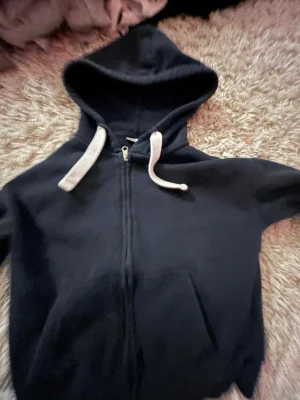 Marinblå hoodie från Gina Tricot - Klassisk marinblå hoodie från Gina Tricot med dragkedja framtill, vit dragsko i huvan och en stor magficka. Tillverkad i mjukt och mysigt material, perfekt för chill dagar. Enkel och stilren design som passar till det mesta. 