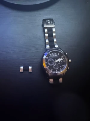 Armani Exchange AX-klocka i silver - Stilren klocka från Armani Exchange med svart urtavla och silverfärgade detaljer. Länken är i metall med svarta inslag och har AX-logga på spännet. Kronograf-funktion och tydliga markeringar på urtavlan. En extra länkbit medföljer. Samt med en batteri. (Lite repor på baksidan)