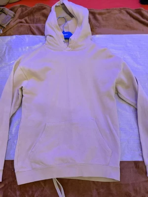 Beige mjukisset med hoodie och byxor - Mysigt loungewear-set i beige med hoodie och matchande byxor. Hoodien har känguruficka och dragsko i huvan, byxorna har resår i midjan och dragsko för skön passform. Perfekt för chill dagar hemma eller när du vill vara extra bekväm.