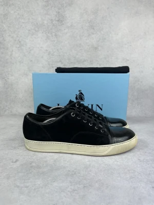 Svarta sneakers från Lanvin - Stilrena svarta sneakers från Lanvin med ovandel i mocka och läderdetaljer på tån och hälen. Skorna har vita gummisulor och svarta skosnören. Perfekta för dig som gillar en clean och klassisk look.