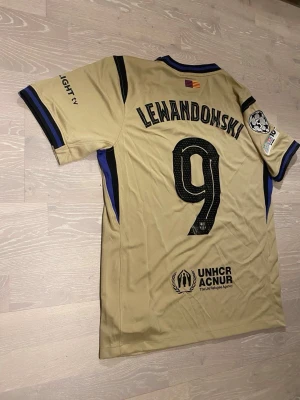 Beige FC Barcelona Lewandowski tröja - Säljer en beige FC Barcelona matchtröja med Lewandowski och nummer 9 på ryggen. Tröjan har svarta och blå detaljer, Champions League-märke på ärmen och UNHCR-logga nedtill. Tillverkad i lätt och ventilerande material, perfekt för fotboll eller träning.