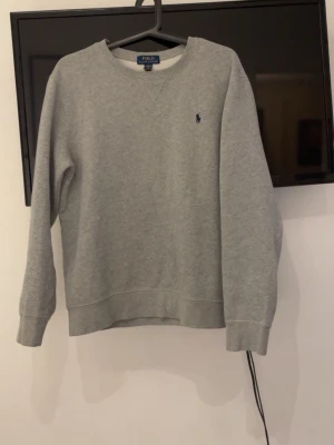 Grå sweatshirt från Polo Ralph Lauren - Klassisk grå sweatshirt från Polo Ralph Lauren med rund halsringning och diskret blå logga broderad på bröstet. Tröjan har ribbade muddar vid ärmslut och nederkant. Perfekt för en avslappnad och stilren look. Storlek 170, men är ungefär som en mindre S