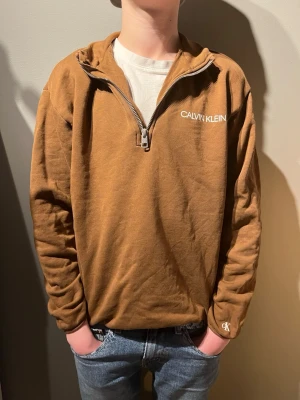 Brun halfzip från Calvin Klein - Snygg brun tröja från Calvin Klein med halv dragkedja och vit logga på bröstet. Tillverkad i mjukt bomullsmaterial och har en relaxed passform. Perfekt till jeans eller över en t-shirt för en avslappnad look.