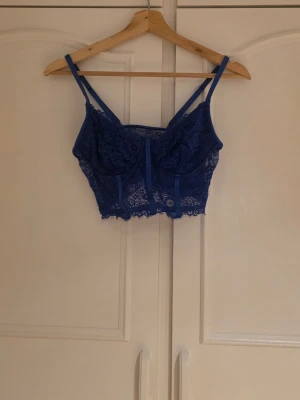 Blå spets bustier med smala band - Snygg blå bustier med tunna axelband och detaljer i spets. Toppen har en croppad passform och genomskinliga spetsdetaljer både fram och bak. Aldrig använd 