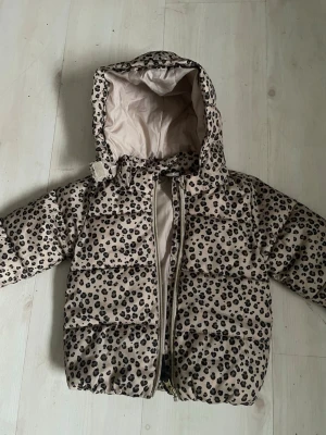 HM Beige pufferjacka med leopardmönster - H M Snygg beige pufferjacka med leopardmönster och huva. Jackan har dragkedja framtill och quiltad design som håller värmen. Perfekt för kalla vinterdagar och ger en trendig look. Materialet är troligtvis polyester.