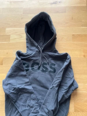 Mörkgrå hoodie från BOSS - Snygg mörkgrå hoodie från BOSS med stor logga på bröstet. Klassisk modell med huva och dragsnören samt en stor magficka framtill. Tillverkad i mjukt bomullsmaterial som känns skönt mot huden. Perfekt för en avslappnad och trendig look.