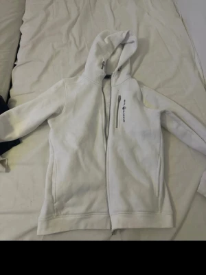 Vit hoodie från Peak Performance - Snygg vit hoodie från Peak Performance med dragkedja framtill och broderad logga på bröstet. Hoodien har en klassisk huva och två fickor på sidorna. Perfekt för en avslappnad och sportig stil.