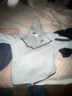 Ljusgrå Nike hoodie med dragkedja - Ljusgrå hoodie från Nike med hel dragkedja och svart Nike-logga på bröstet. Tröjan har huva, svarta detaljer längs dragkedjan och muddar vid ärmslut och nederkant. Perfekt för en sportig och avslappnad stil.