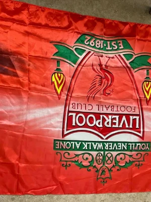 Liverpool FC röd supporterflagga - Stor röd flagga med Liverpool FC:s klassiska klubbmärke, gröna och gula detaljer samt texten 'You'll Never Walk Alone'. Perfekt för att visa sitt stöd för laget på matcher eller hemma.