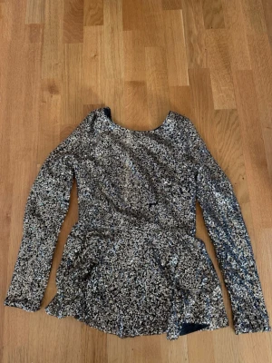 Glittrig långärmad topp med öppen rygg - Säljer en glittrig guldfärgad topp med paljetter, lång ärm och snygg öppen rygg. Toppen har en figurnära passform och är täckt av paljetter som ger ett riktigt statement-utseende. Perfekt för dig som vill sticka ut och glänsa.
