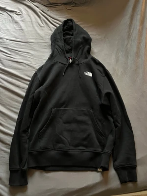 Svart hoodie från The North Face - Svart hoodie från The North Face med vit logga på bröstet och stort tryck på ryggen med texten 'Never Stop Exploring'. Klassisk känguruficka framtill, ribbade muddar och justerbar huva. Perfekt för en avslappnad streetwear-look. Kom gärna med prisförslag. 