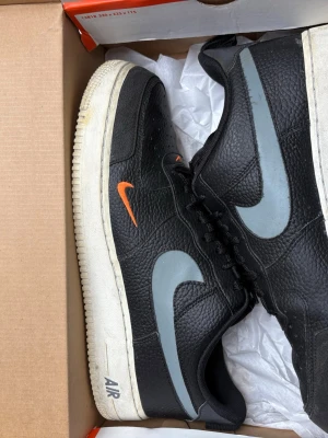 Nike Air Force 1 svart med grå swoosh - Svarta Nike Air Force 1 sneakers i läder med grå swoosh på sidorna och en liten orange swoosh på utsidan. Klassisk vit sula med 'AIR'-text på sidan. Lågt skaft och snörning framtill. Perfekta för en streetwear-look.
