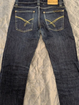 Mörkblå jeans från Deminbirds - Snygga mörkblå jeans från Deminbirds med raka ben och kontrastsömmar i gult. Bakfickorna har dekorativa broderier och klassisk femficksdesign. Jeansen är tillverkade i ren denim och har en tidlös look som passar till det mesta.
