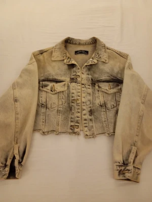 Cropped ljusgrå jeansjacka - Trendig cropped jeansjacka från dilvin jeans i ljusgrå tvätt med slitna detaljer och rå kant nertill. Klassisk krage, två bröstfickor med lock och knapp samt knappar vid ärmslut. Perfekt för en edgy streetstyle-look.