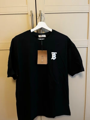 Svart Burberry t-shirt storlek M/L - Snygg svart t-shirt från Burberry med vit TB-logga på bröstet. Klassisk rund halsringning och normal passform. Perfekt för dig som vill ha en stilren och trendig look med exklusiv känsla. Tillverkad i mjuk bomull för maximal komfort.