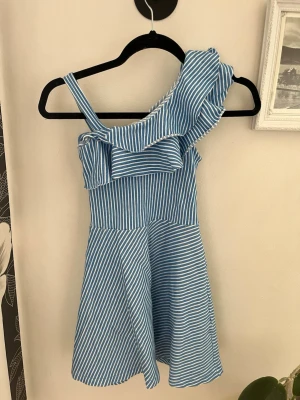 Barn Blåvit randig oneshoulder klänning - Snygg blå och vit randig kortklänning med volangdetalj och oneshoulder-design. Klänningen har en figurnära överdel och utställd kjol, perfekt för varma dagar. Tillverkad i ett lätt och bekvämt material.Barn stl 134
