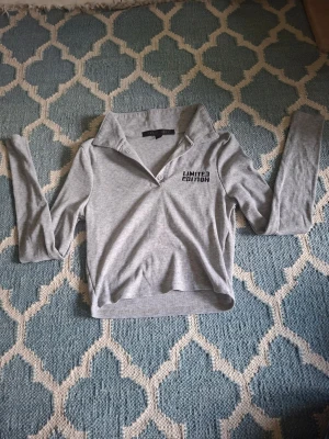 Grå ribbad långärmad topp - Grå ribbad långärmad topp från Bershka med krage och knapp framtill. Trycket 'LIMITED EDITION' på bröstet ger en cool vibe. Toppen har en croppad passform och är perfekt att styla med jeans eller kjol.