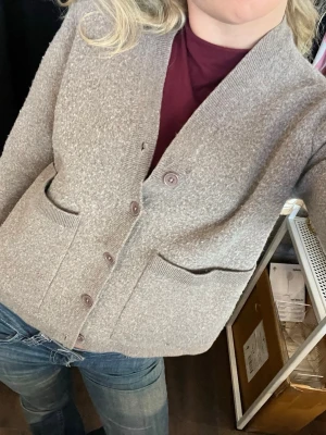 Snygg Beige kofta från A Day’s March - Snygg beige / ljusbrun kofta från a days march  med v-ringning och knappar framtill. Koftan har två stora fickor och är tillverkad i ett mjukt, stickat 100% lamm ull material. Perfekt för lager på lager och enkel att matcha med olika outfits.