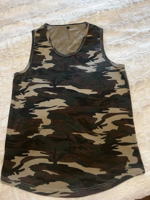 Camouflagefärgad linnetopp - Säljer en ärmlös linnetopp i klassiskt camouflage-mönster med gröna, bruna och beige toner. Toppen har rund halsringning och är perfekt för dig som gillar en avslappnad och cool stil. Materialet känns mjukt och lätt mot huden.