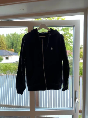 Svart hoodie RP - Svart hoodie med dragkedja. Klassisk design med vit broderad logga på bröstet, huva med dragsko och två fickor framtill. Perfekt för en chill och stilren look.                  Det är storlek M men passar också S.