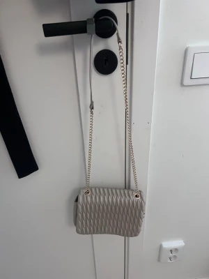 Beige axelväska med kedjerem - Snygg beige axelväska med quiltat mönster och guldfärgad kedjerem. Väskan har en mjuk, rundad form och stängs med lock. Perfekt för dig som vill ha en stilren och trendig accessoar till din outfit.
