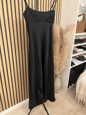 Svart lång klänning med smala axelband - Stilren svart lång klänning med smala axelband och lätt utsvängd passform. Klänningen har en enkel och elegant design, perfekt för dig som gillar minimalistisk stil. Materialet har en lätt glansig yta som ger en lyxig känsla. Skriv om ni vill ha bild på 🫶🏼🫶🏼