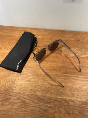 Solglasögon från Prestige - Snygga grå solglasögon från Prestige med transparent båge och mörka glas. Medföljer en svart tygpåse. 