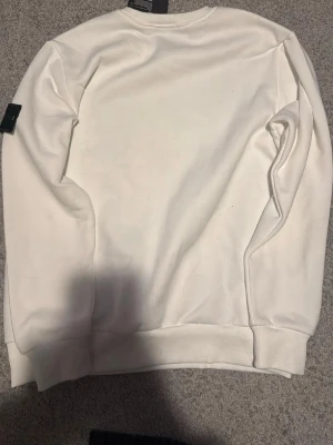 Vit sweatshirt från Stone Island - Vit sweatshirt från Stone Island med klassisk rund hals och ribbade muddar. Tröjan har den ikoniska svarta logotyp-patchen med gult och grönt broderi på vänster ärm. Perfekt för en clean och stilren look.
