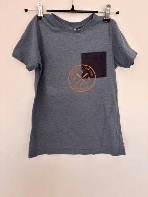 Blå t-shirt Pompdeluxe, strlk 128 - Snygg blå t-shirt med svart bröstficka och orange kompass-tryck på framsidan. T-shirten har rund hals och korta ärmar, perfekt för en avslappnad stil. Materialet känns mjukt och bekvämt, troligen bomull.