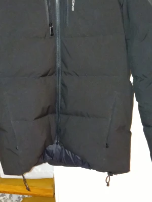 Svart dunjacka från Everest - Snygg svart dunjacka från Everest med dragkedja framtill och två sidofickor. Jackan är vadderad med 500 FP dun och har justerbar dragsko nedtill. Perfekt för kalla vinterdagar när du vill hålla dig varm och stilren.