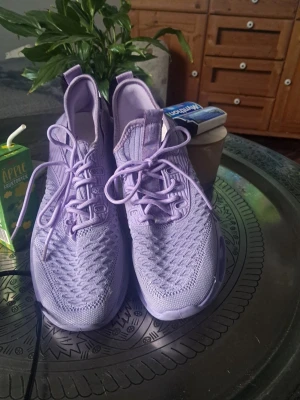 Lila stickade sneakers med snörning - Snygga sneakers i en trendig lila färg med stickad ovandel och matchande lila snören. Skorna har en sportig design med låg profil och ventilerande meshmaterial som ger en modern look. Perfekta för dig som vill sticka ut med färg på fötterna.
