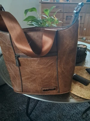 Brun bucket bag i fejkläder - Snygg brun bucket bag i fejkläder med grov struktur och breda axelband i textil. Väskan har en praktisk ytterficka med dragkedja och en stilren, modern look. Perfekt för dig som vill ha en rymlig och trendig handväska.