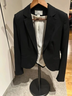Svart kavaj från H&M, storlek 36 - Stilren svart kavaj från H&M i storlek 36. Klassisk modell med smal passform, spetsiga slag och två fickor framtill. Fodret är ljus med tunna ränder. Perfekt för en snygg och uppklädd look.  Se bild med H&M loggan där det är en missfärgning, i övrigt gott skick.