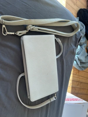 Ljusgrå kuvertväska med axelrem - Stilren ljusgrå kuvertväska i rektangulär form med avtagbar och justerbar axelrem. Väskan har en diskret struktur i materialet och enkel design utan synliga loggor. Perfekt för att bära det viktigaste med en clean look.