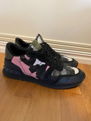 Valentino sneakers med camomönster - Snygga sneakers från Valentino med svart, grått, vitt och rosa camomönster. Skorna har svarta snören, svarta detaljer i mocka och läder samt en cool logga på plösen. Perfekta för dig som vill sticka ut med en unik och trendig stil. På bild 5 har det där fram lossat lite