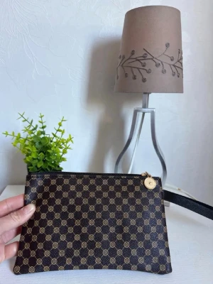 Brun mönstrad Kuvertväska   - Snygg brun kuvertväska med klassiskt mönster i beige och brunt.Plånboken har dragkedja med guldfärgad detalj och en praktisk handledsrem. Perfekt för att hålla ordning på kort och småsaker. Materialet är tåligt och lätt att torka av.