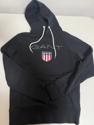Svart hoodie från GANT med logga - Svart hoodie från GANT med vit dragsko i huvan och broderad GANT-logga samt sköld med stjärnor och ränder på bröstet. Klassisk känguruficka framtill och ribbade muddar. Perfekt för en avslappnad och stilren look.