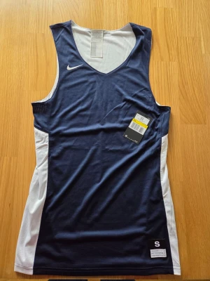 Nike träningstop , basket , NBA, strorlek S - Sportigt basketlinne från Nike i marinblått med vita sidopaneler. Tillverkat i lätt och ventilerande meshmaterial som passar perfekt för träning och matcher. Klassisk V-ringning och broderad Nike-logga på bröstet. Snygg och funktionell design.
