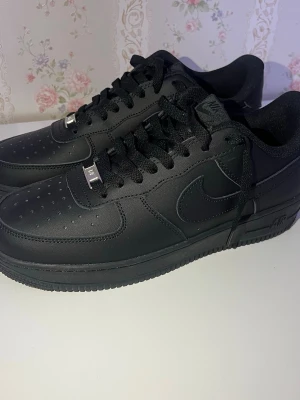 Nike Air Force 1 helsvarta sneakers - Klassiska Nike Air Force 1 sneakers i helsvart läder med svart sula och svarta skosnören. Ikonisk Swoosh-logga på sidorna och perforerad tå för extra ventilation. Perfekta för en clean och stilren look.