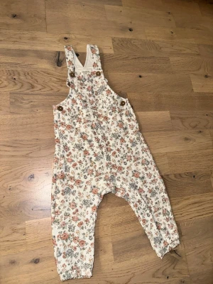 Blommig byxdress från Newbie - Supersöt byxdress från Newbie i storlek 74 med blommigt mönster i pastellfärger. Byxdressen har axelband med knappar och resår vid bensluten. Perfekt för dig som gillar romantisk och lekfull stil.