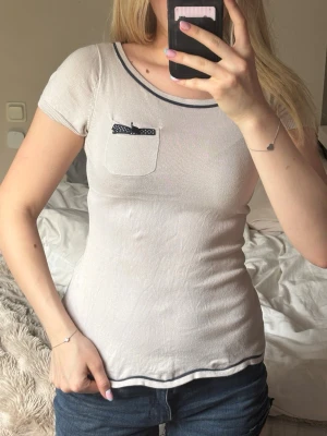 Rosa topp med rosett och ficka - Söt rosa topp med korta ärmar och rund halsringning. Svarta detaljer vid hals och fåll samt en liten bröstficka med svartvit prickig rosett. 
