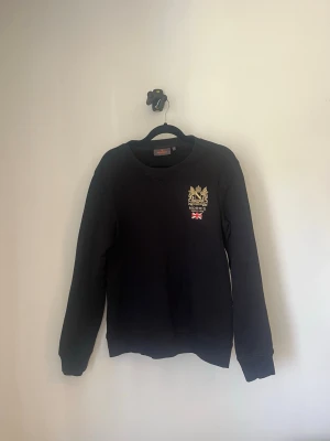 Svart stickad tröja från Morris - Svart sweatshirt från Morris med rund halsringning och broderad logga. Perfekt för en stilren och klassisk look. Storlek S, Använd fåtal gånger skick 7/10
