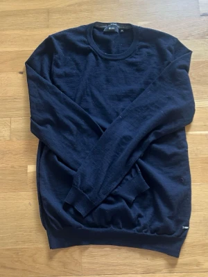 Mörkblå stickad tröja från BOSS - Snygg mörkblå stickad tröja från BOSS med rund halsringning och diskret metallogo vid nederkanten. Tröjan har långa ärmar och ribbade muddar vid ärmslut och midja. Perfekt för en stilren och klassisk look. Merinoull 