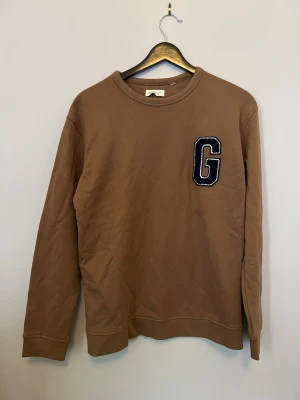 Brun sweatshirt från Gant med G-logga - Säljer en brun sweatshirt från Gant med en stor blåvit G-logga på bröstet. Tröjan har rund halsringning, ribbade muddar och är tillverkad i mjukt bomullsmaterial. Perfekt för en avslappnad och stilren look.