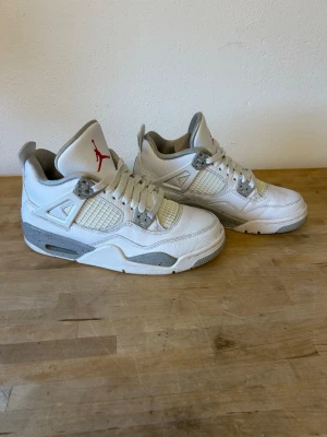 Air Jordan 4 Retro White Oreo sneakers  - Säljer ett par Air Jordan 4 White Oreo sneakers i vitt läder med grå och svarta detaljer samt röd Jumpman-logga på plösen. Klassisk siluett med meshpaneler och robust sula. Använda men i fint skick. Orginalsnören och creasingskydd ingår, Obs! Ingen orginalbox. Storlek 38.