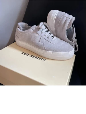 Beige sneakers från Axel Arigato - Snygga beige sneakers från Axel Arigato i mocka med klassisk låg siluett. Skorna har vita snören och en tjock vit sula som ger en clean och stilren look. Perfekta för dig som gillar minimalistisk design och premiumkänsla.